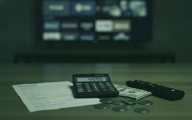 A calculator, dollar bills, coins, a streaming stick and a remote on a table in front of a TV